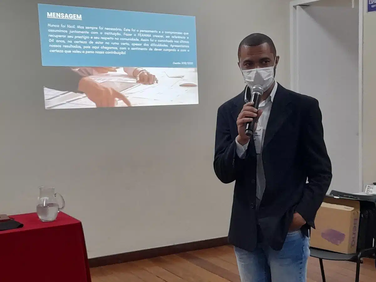 Júnior Eduardo Nonato, usando máscara protetora, fala ao microfone durante a apresenta a prestação de contas da Federação das Associações de Moradores de Mariana, durante o segundo congresso, tendo ao fundo a projeção de mensagem aos participantes