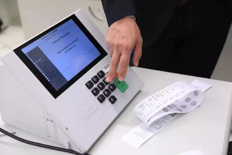Urna eleitoral, sobre uma mesa, em teste feito, observando-se uma mão masculina pressionando uma das teclas e uma fita de papel com boletim da urna