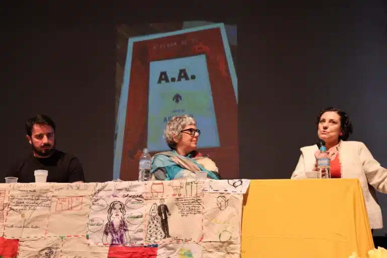 Setembro amarelo: Debatedores em uma mesa com toalha amarela, à frente de um fundo escuro, com a projeção da capa de um livro de psicologia