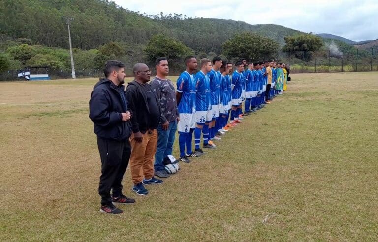 Jogadores, dirigentes e trio de arbitragem formam uma fila no centro do campo, durante a execução dos hinos na cerimônia de abertura da final do campeonato de juniores de Mariana