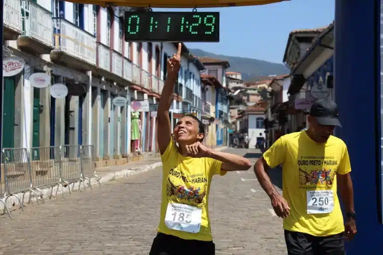 Deficiente visual, usando camiseta amarela e short preto, cruza a linha de chegada na Rua Direita, ao final da 9ª Corrida Mariana - Ouro Preto