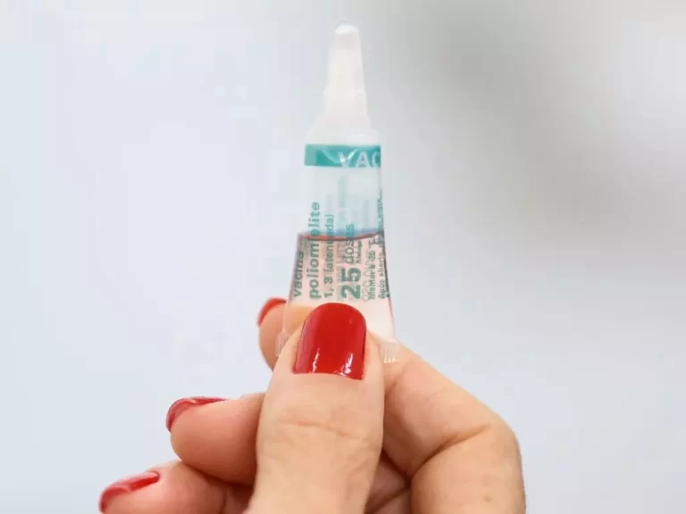 Mão de mulher, com unhas pintadas de vermelho, segura um tubo de vacina contra a poliomielite