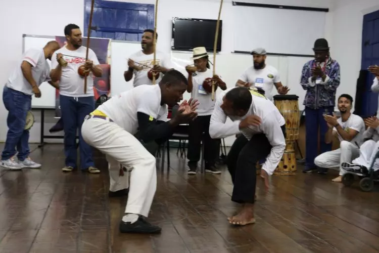 Dois homens jogam capoeira em uma sala. Ao fundo, outros participantes tocam percussão e berimbau. A atividade da capoeira também foi incentivada com emendas impositivas