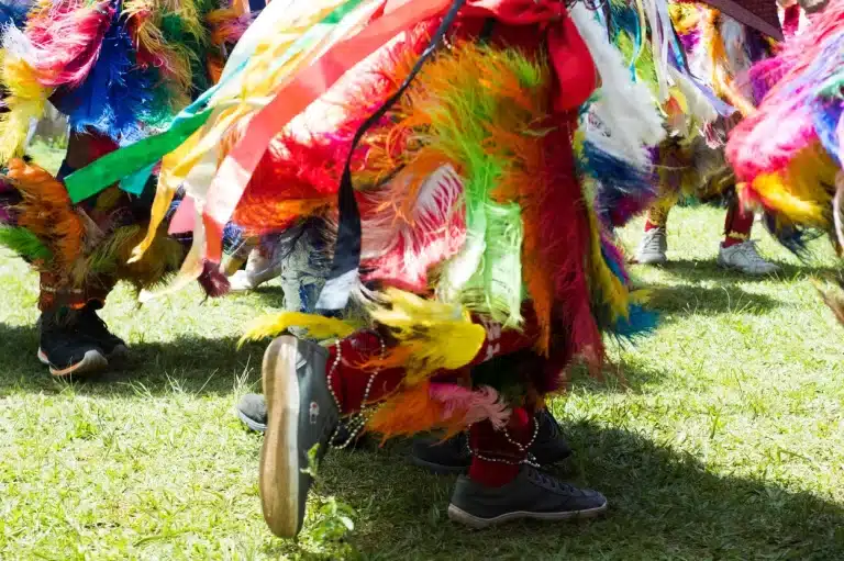 Sobre um gramado, pessoas dançam, usando roupas com penas e fitas multicoloridas