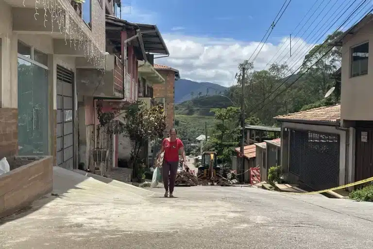 Mulher com blusa vermelha e calça azul sobre a Rua João Batista, em Passagem de Mariana, carregando sacolas, tendo ao fundo o equipamento utilizado para executar a obra de reparo da via.