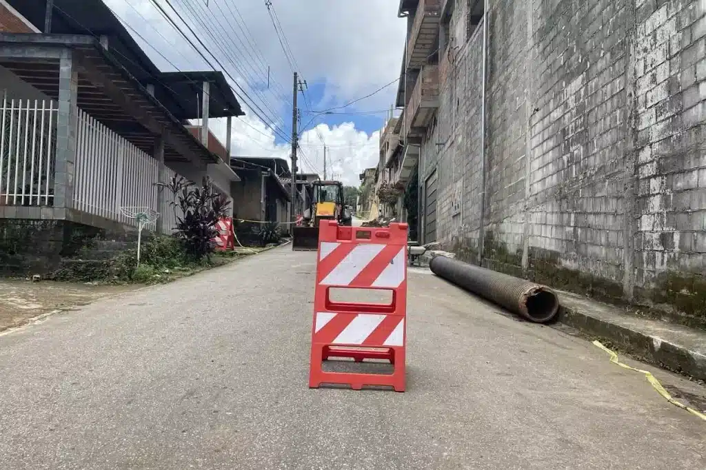 Cavalete em vermelho e branco sinaliza a interdição da rua João Batista, em Passagem de mariana. Ao fundo, tubulação para reparo de rede pluvial e trator para a execução do serviço